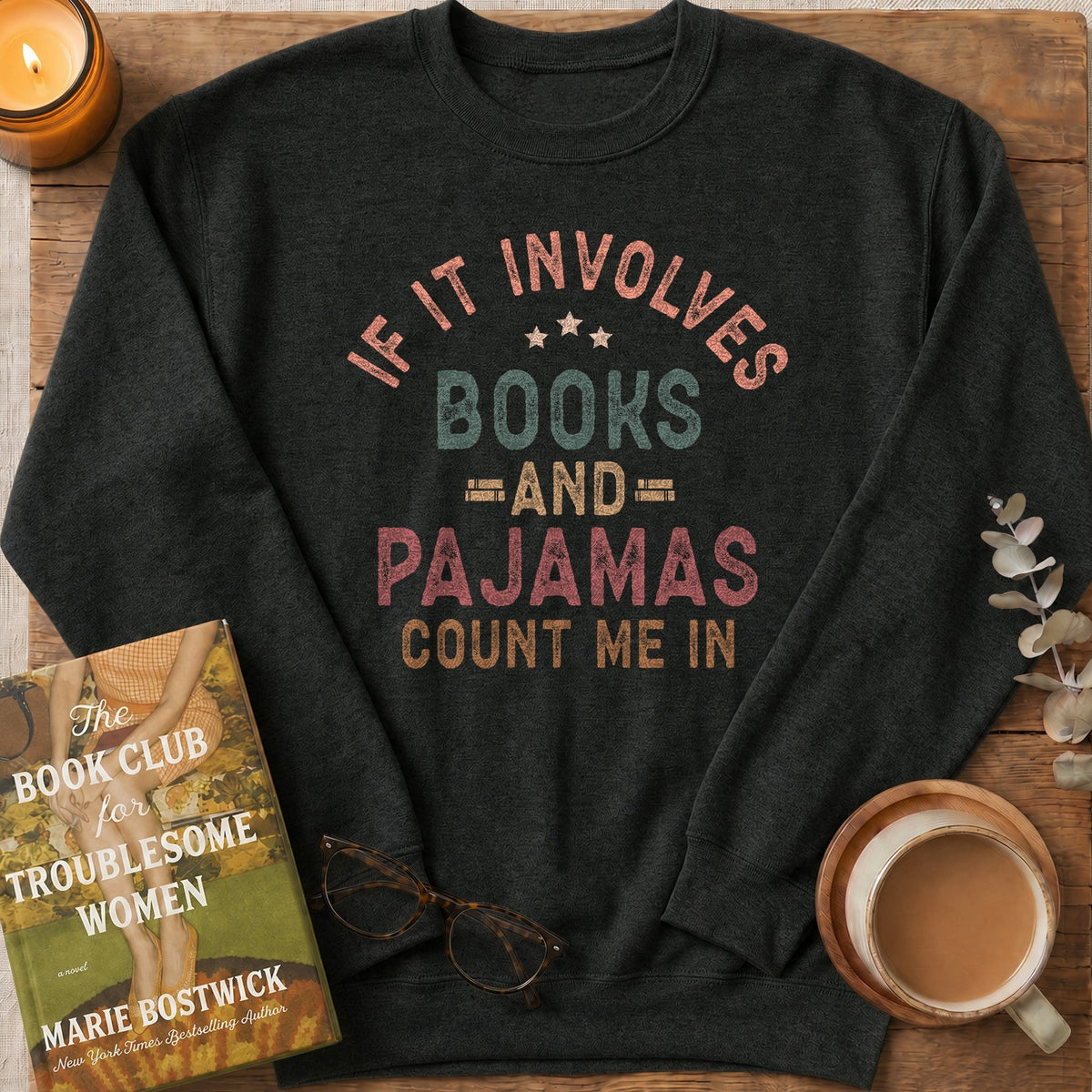 A dark charcoal gray long-sleeve t-shirt with a retro-style graphic reading 'IF IT INVOLVES BOOKS AND PAJAMAS COUNT ME IN' in muted pink, teal, and tan lettering. The shirt is styled in a cozy flat-lay arrangement on a rustic wooden board, surrounded by a lit candle, a book titled 'The Book Club for a Troublesome Women' by Marie Bostwick, eyeglasses, and a cup of coffee.