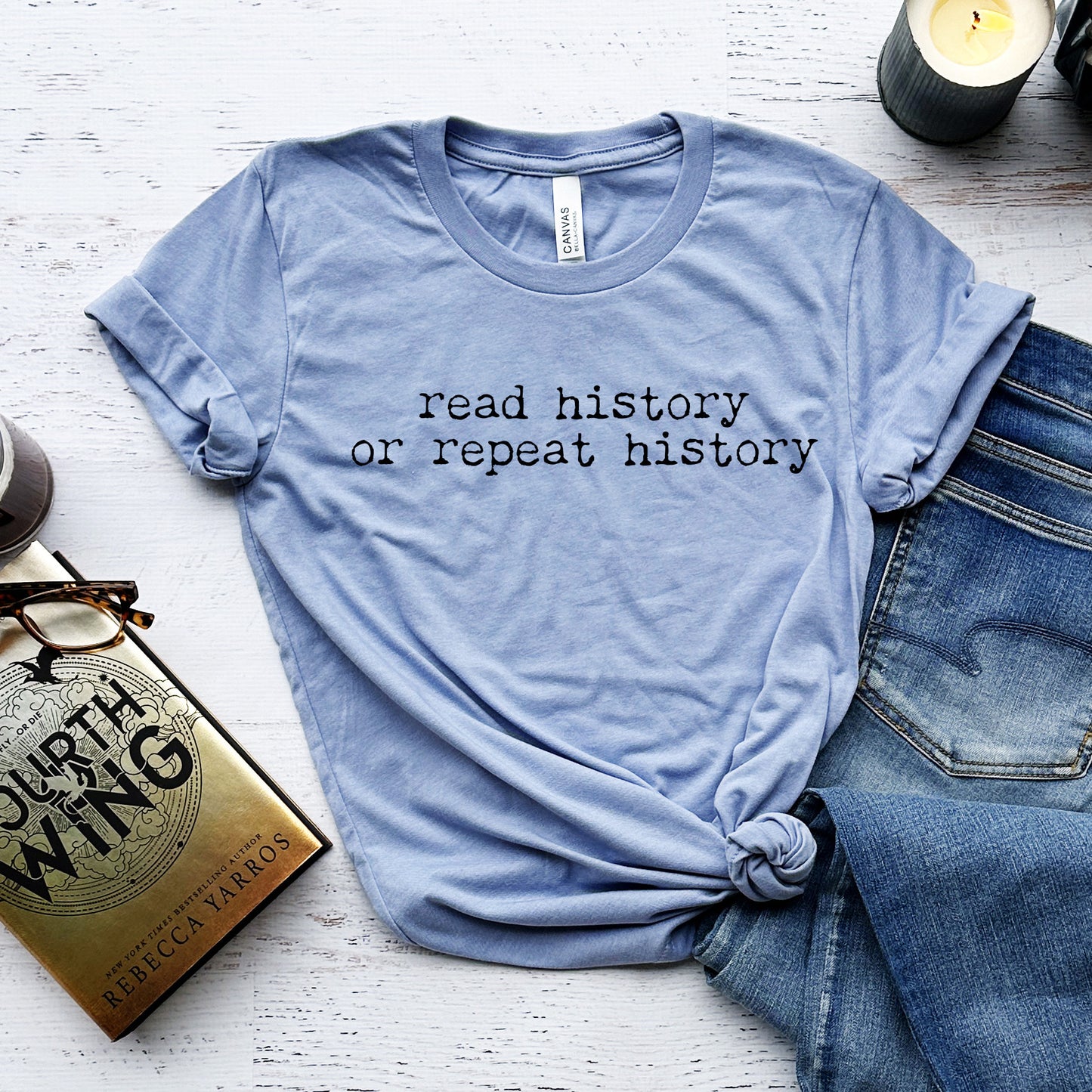 Blue t-shirt with 'read history or repeat history' text on a white surface with the book Fourth Wing by Rebecca Yarros and jeans.