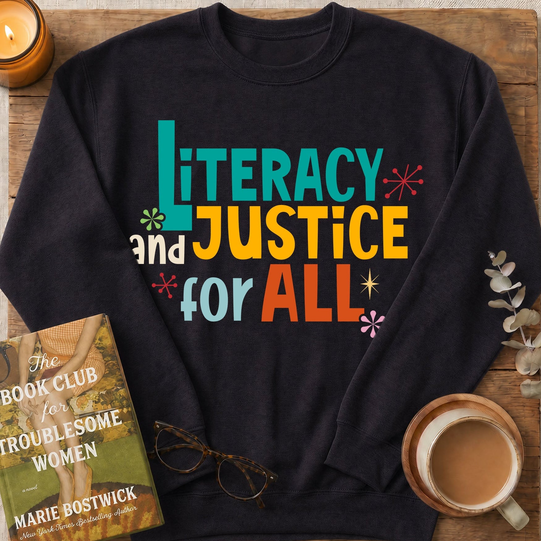 A black crewneck sweatshirt with a colorful graphic reading 'Literacy and Justice for All' in teal, yellow, orange, and white. The sweatshirt is presented in a styled flat-lay on a wooden board with a book, glasses, a cup of coffee, a candle, and eucalyptus branches.