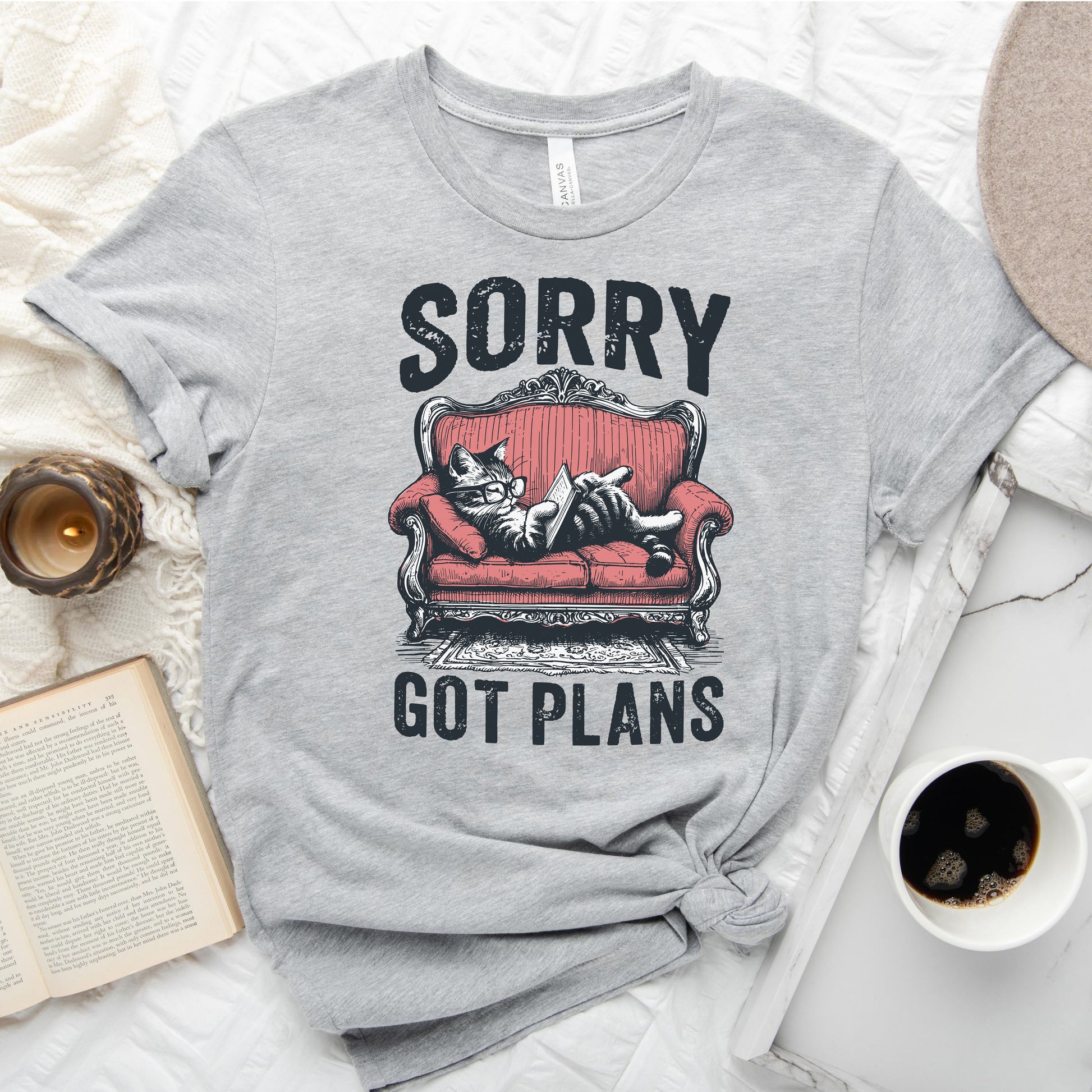 Gray t-shirt with a graphic of a cat on a red couch and text 'Sorry, got plans' on a white surface with a book and coffee cup.