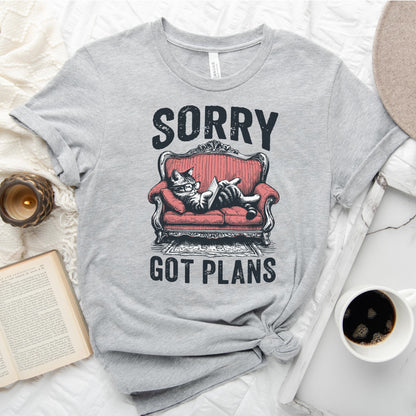 Gray t-shirt with a graphic of a cat on a red couch and text 'Sorry, got plans' on a white surface with a book and coffee cup.