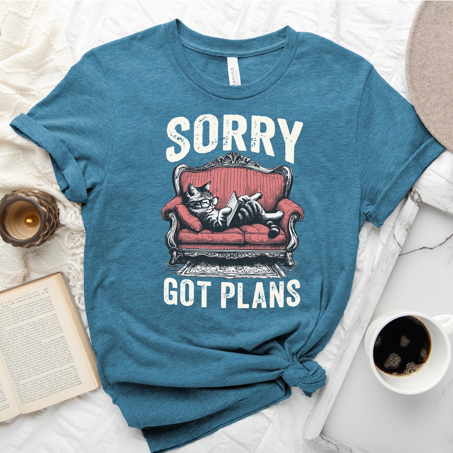 Blue t-shirt with a graphic of a cat on a red couch and text 'Sorry, got plans' on a white surface with a book and coffee cup.