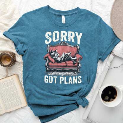 Blue t-shirt with a graphic of a cat on a red couch and text 'Sorry, got plans' on a white surface with a book and coffee cup.