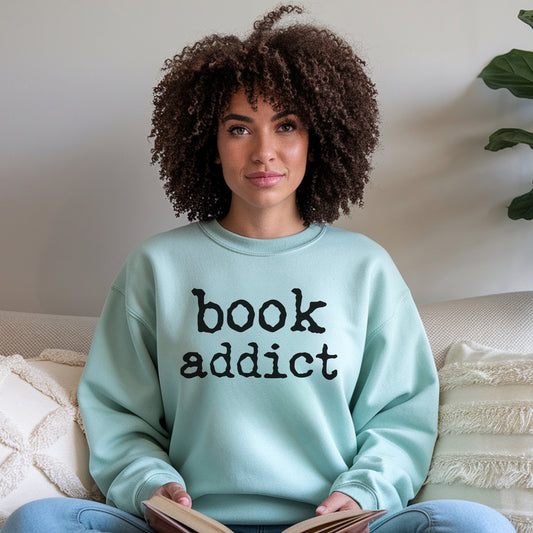 Woman wearing a light blue sweatshirt with 'book addict' text, sitting on a couch holding a book.