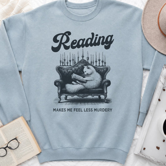 A light blue crewneck sweatshirt laid flat on a white fabric background, featuring a black graphic of a cat reading on a chaise lounge with candles, and the text 'Reading MAKES ME FEEL LESS MURDERY'. A book, glasses, coffee cup, and hat are styled around it as props.