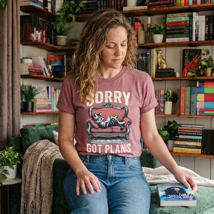 Woman sitting on a couch wearing a t-shirt with a vintage cat on a couch reading a book with the text Sorry got plans, in a room with bookshelves and plants.